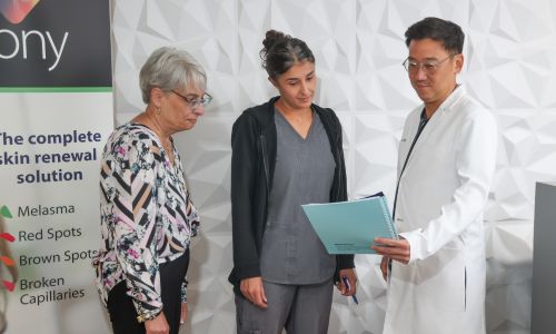 A doctor in a white coat reviews a file with two women, one in casual wear and the other in medical scrubs, in a clinic setting with a poster about skin treatments in the background.