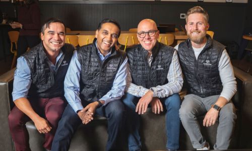 Four men sitting on a couch, all wearing casual attire and matching black vests, smiling at the camera. The background includes a dark wall and yellow chairs.