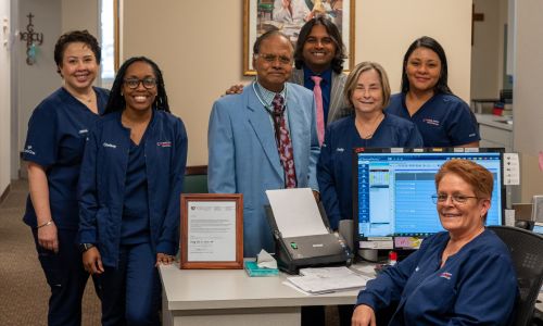 A group of six healthcare professionals, five women in navy uniforms and one man in a suit, stand and smile behind a desk in an office setting. A computer and various office supplies are on the desk.