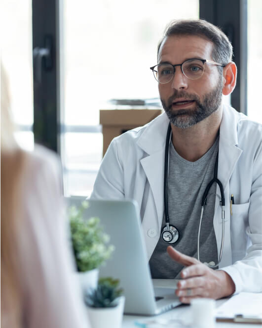 A doctor wearing a white coat and stethoscope, sits at a desk with a laptop, talking to a patient.