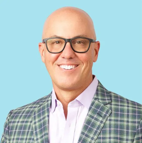 Bald man with glasses wearing a plaid jacket and light purple shirt smiles against a light blue background.