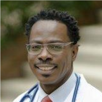 A man with short curly hair, wearing glasses, a white coat, and a red tie, smiling outdoors.