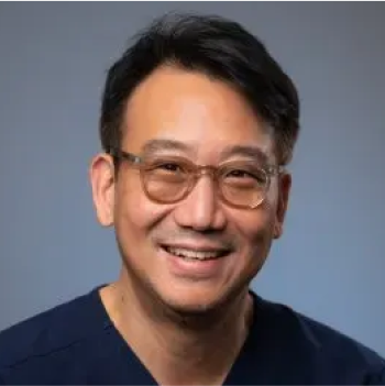 A man with short black hair, wearing glasses and a dark shirt, smiles at the camera against a plain background.