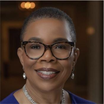 A person with short hair and glasses, dressed in a blue outfit and wearing jewelry, smiles in a warmly lit setting.