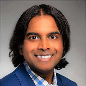 Portrait of a person with dark shoulder-length hair, wearing a blue suit and checkered shirt, smiling at the camera.