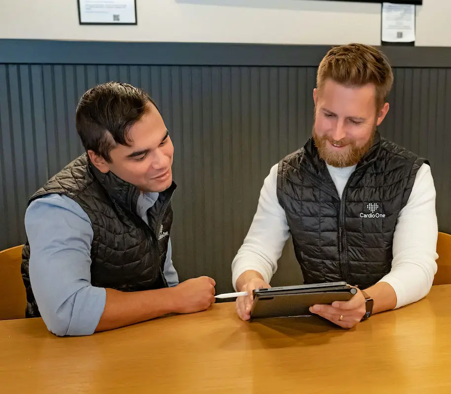 Two men wearing matching black vests sit at a table, smiling and discussing something on a tablet. One man, an independent cardiology physician, holds a stylus while the other leans in to look at the screen.