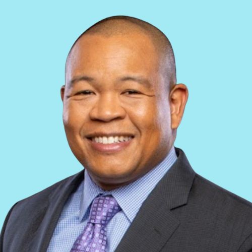 A man in a suit and tie smiles at the camera against a light blue background.