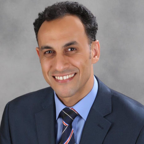 A man with short dark hair, wearing a blue suit, blue dress shirt, and striped tie, smiling in front of a gray background. He embodies the essence of preventative healthcare, representing Houston's dedicated cardiovascular specialists.