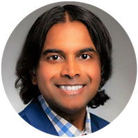 A person with medium-length dark hair wearing a blue suit jacket over a checkered shirt, smiling at the camera in a professional headshot.