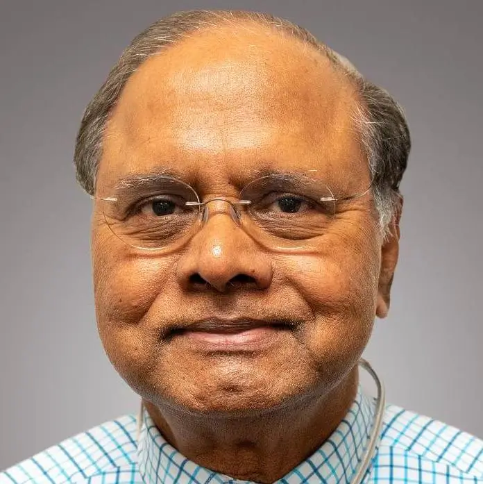 A man with glasses and a checkered shirt smiles at the camera, appearing confident as he stands against a gray background, showcasing his dedication to cardiac prevention.