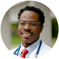 Smiling physician wearing glasses, a white coat, stethoscope, and a red tie, standing outdoors.