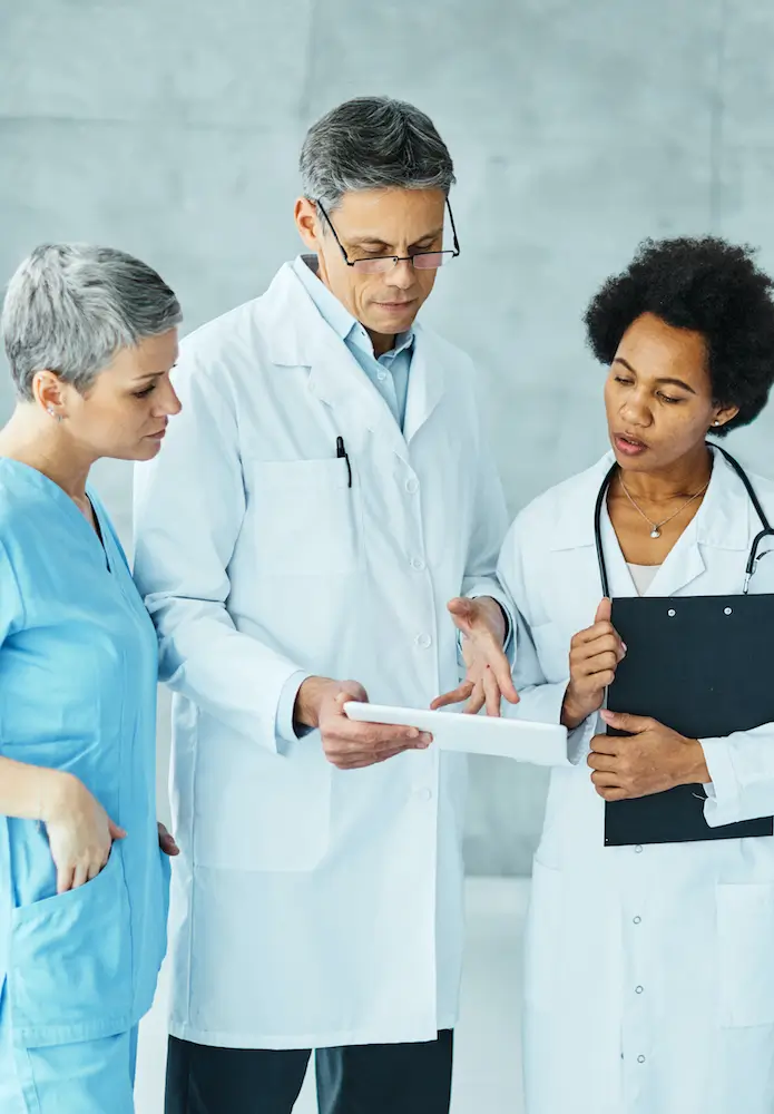 Three medical professionals, including two women and one man, gather in their independent practice to discuss a document. One woman is holding a clipboard as they review best practices.