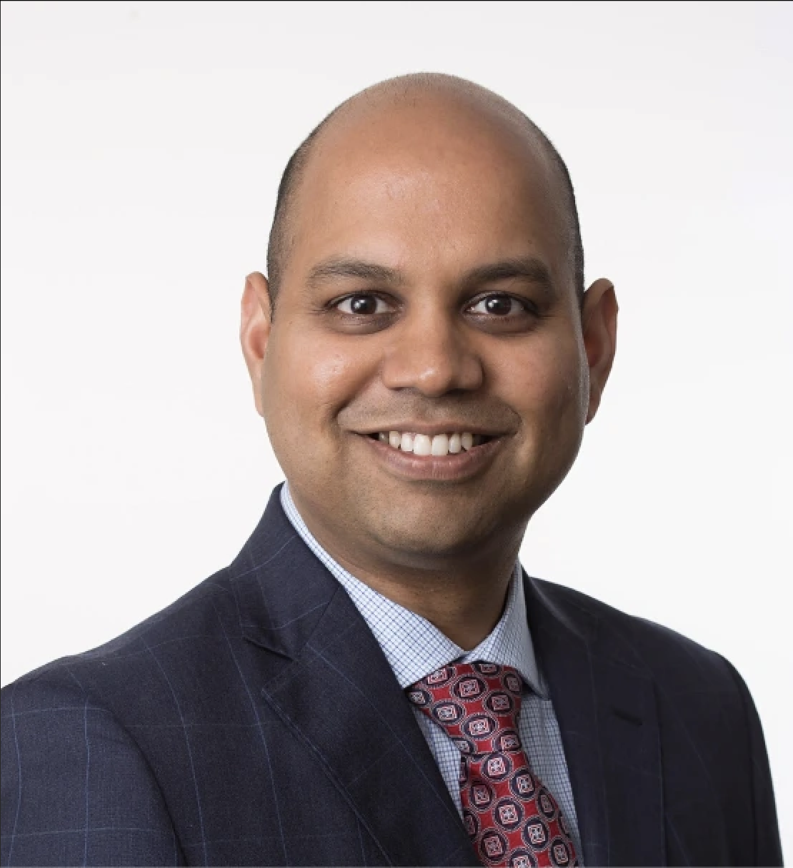 A person wearing a dark suit, light checkered shirt, and patterned red tie is smiling against a plain white background, embodying the look of someone committed to healthy living.