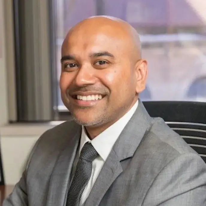 A suited man smiles while seated in an office with a window overlooking Philadelphia in the background.