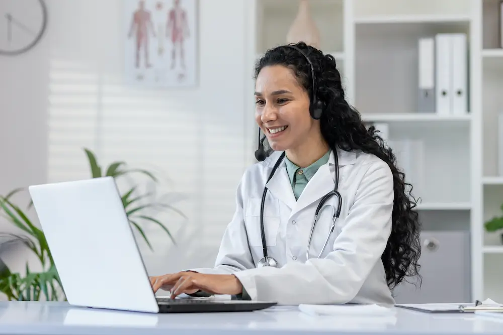 A cardiologist wearing a lab coat and stethoscope sits at a desk, smiling and looking at a laptop while using a headset, likely discussing business planning for their independent practice.