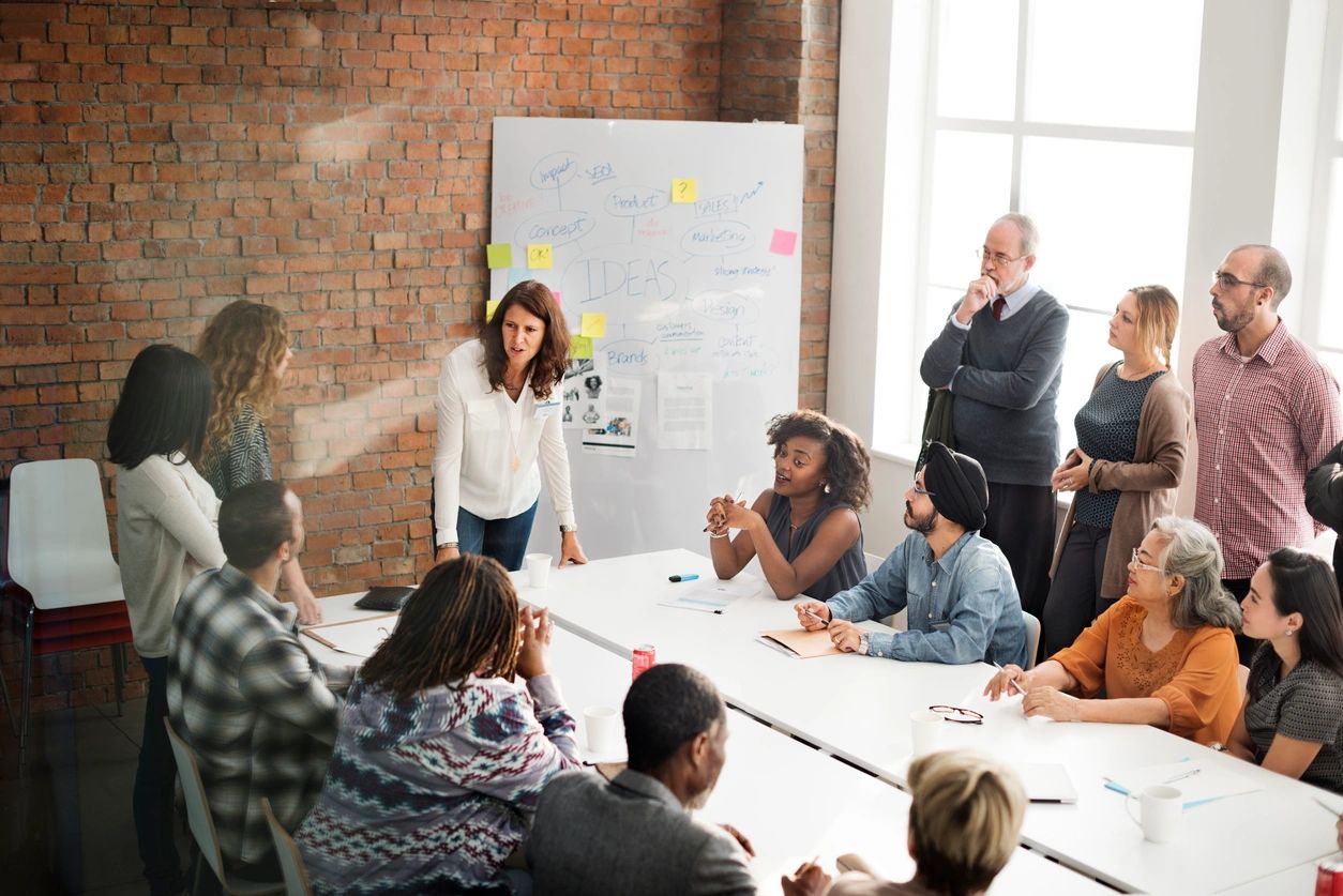 People gathered in a modern office meeting, discussing ideas. A person presents in front of a whiteboard with sketches and notes.