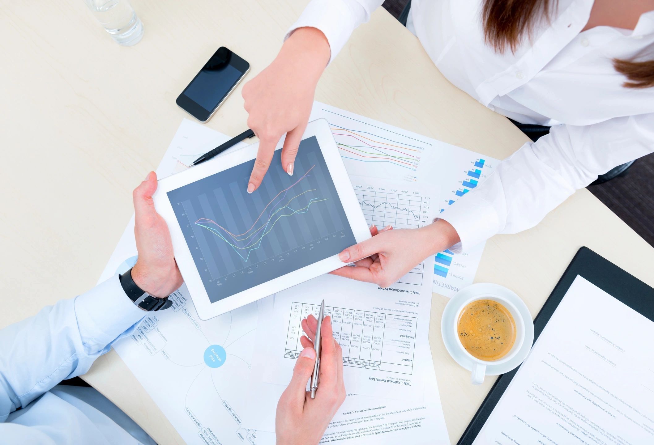 Two people discussing data on a tablet, surrounded by graphs, documents, a smartphone, and a cup of coffee on a table.