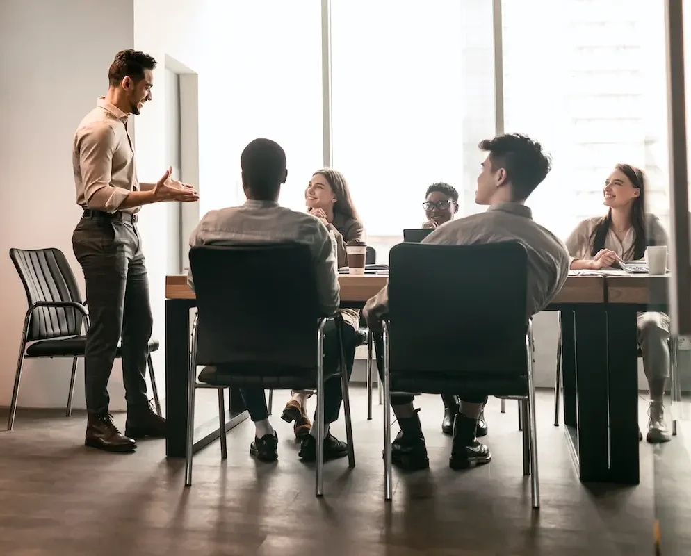 A presenter speaks to a group of coworkers seated around a conference table in a bright, modern office.