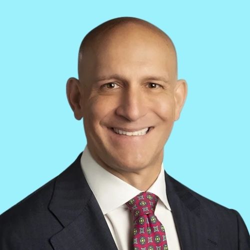 Smiling bald man in a dark suit with a white shirt and pink patterned tie, against a light blue background.
